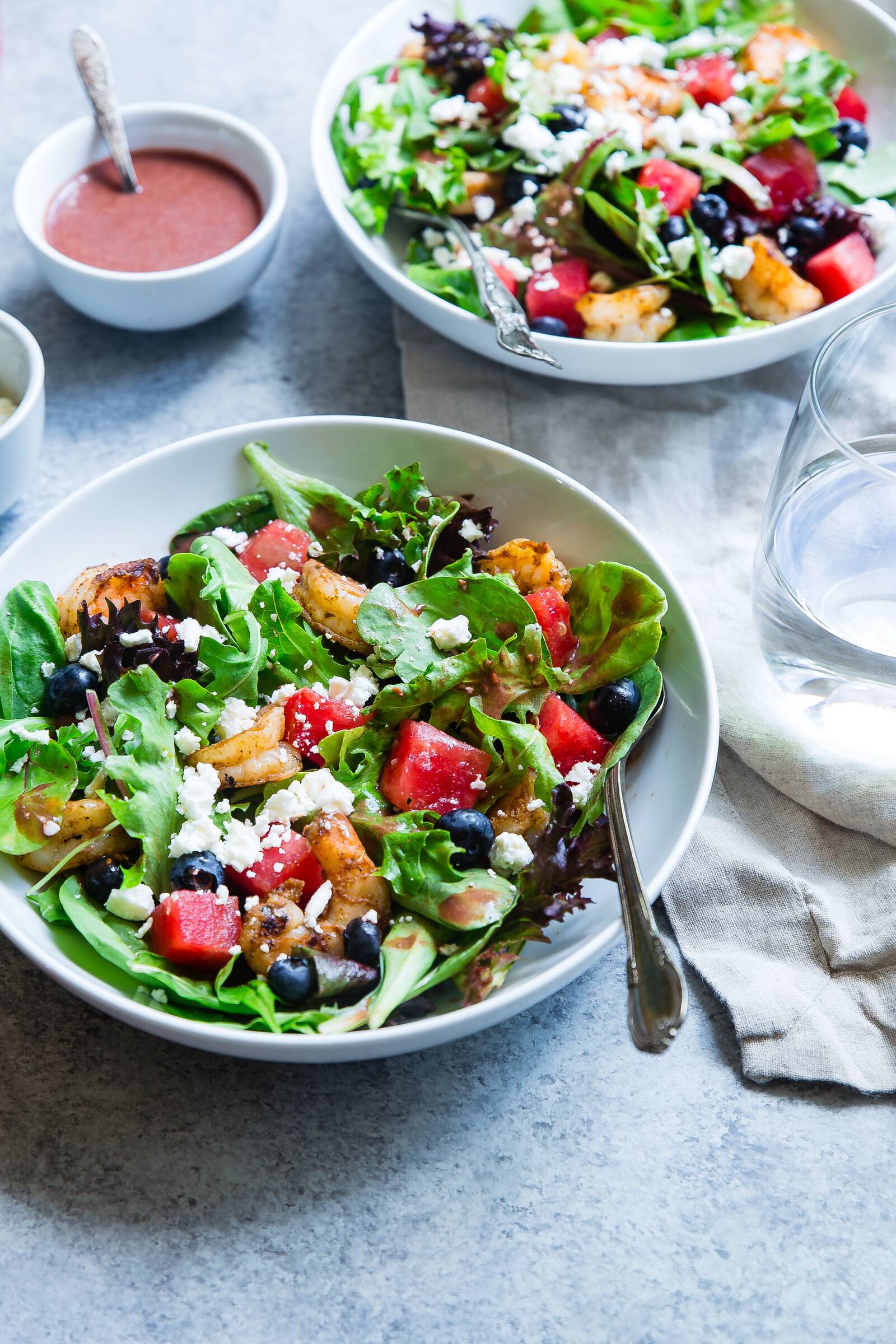 Two bowls of mixed greens salad with grilled shrimp, watermelon, blueberries, and feta, with vinaigrette.