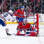 Hockey forward in white reaches for a rebound as red-clad goalie sprawls to block the puck at the net.