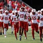 College football team in white and red uniforms runs onto the field as fans cheer in stands.