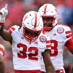 Two Nebraska Cornhuskers football players in white and red uniforms celebrate, No. 32 pointing upward.